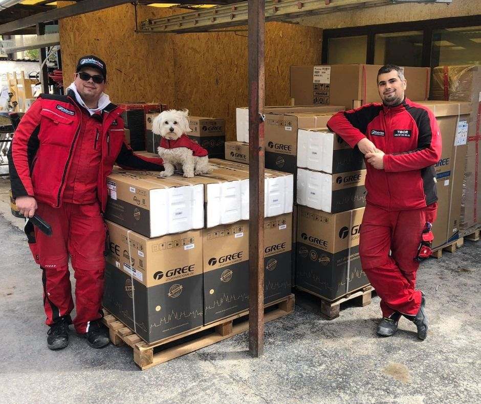 A Valerian Klíma két szerelője piros munkaruhában, egy kis fehér kutyával, raklapon álló Gree klímadobozok között